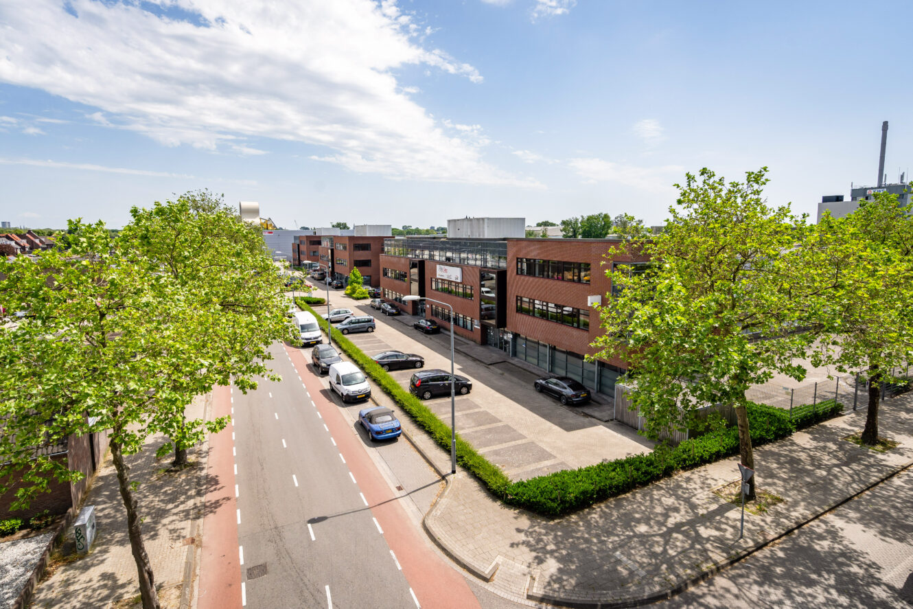 Straatbeeld van een modern kantoorgebouw met geparkeerde auto's, bomen langs het trottoir en een heldere lucht boven je hoofd.