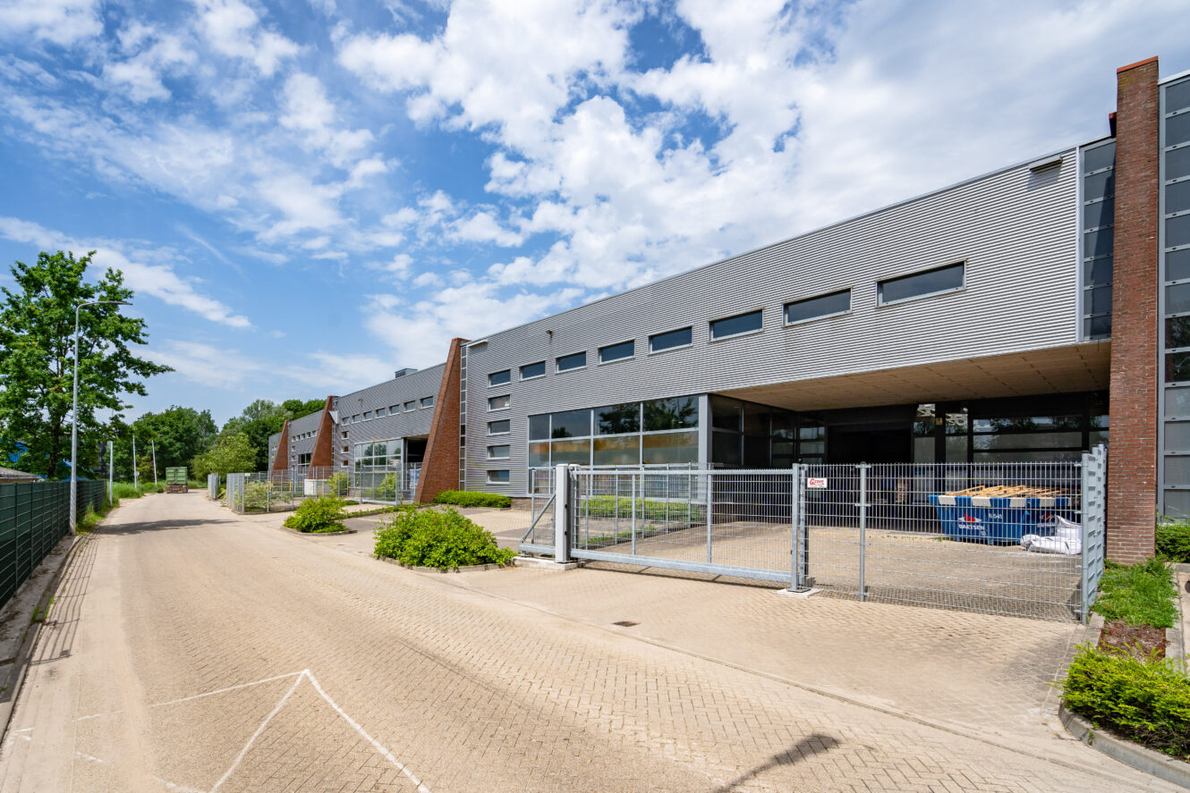 Modern industrieel gebouw met grote ramen en metalen gevelbekleding, poortingang en blauwe lucht met verspreide wolken. Langs het gebouw loopt een verharde weg.