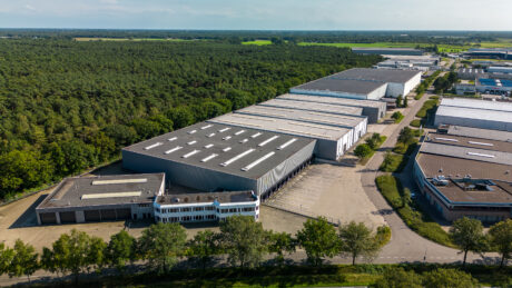 Luchtfoto van een groot industrieel magazijncomplex omringd door bomen, met parkeerplaatsen en aangrenzende gebouwen onder een heldere hemel.