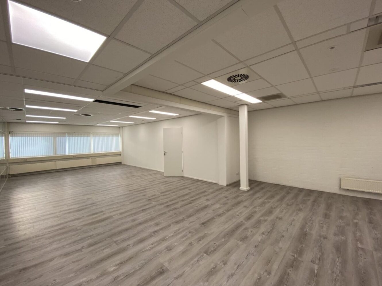 Een ruime, lege kamer met een lichtgrijze houten vloer, witte muren, een witte deur en plafondpanelen met tl-verlichting.