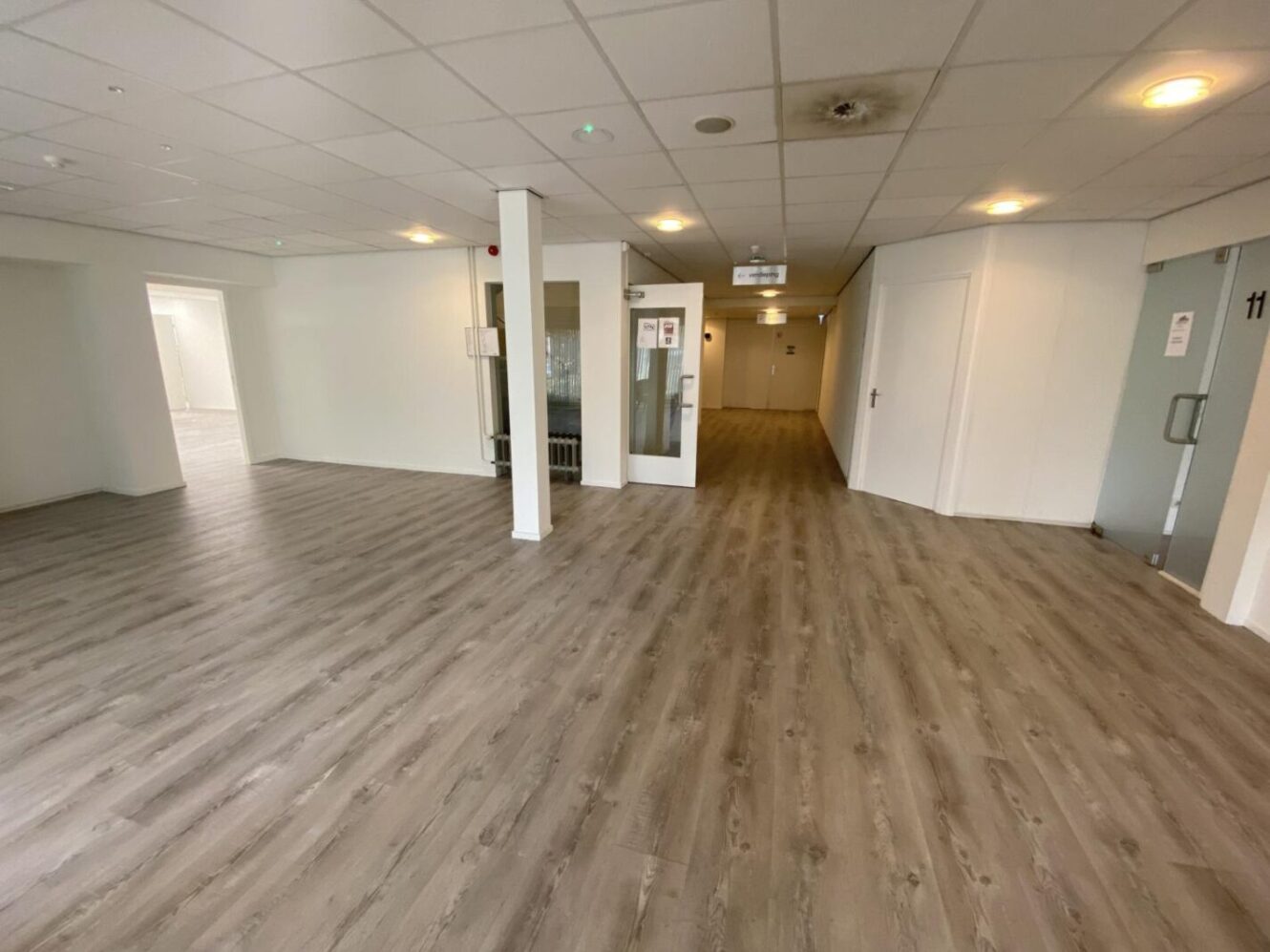 Een grote, lege kamer met lichte houten vloeren, witte muren, een verlaagd plafond met inbouwspots en verschillende deuropeningen die naar andere kamers leiden.