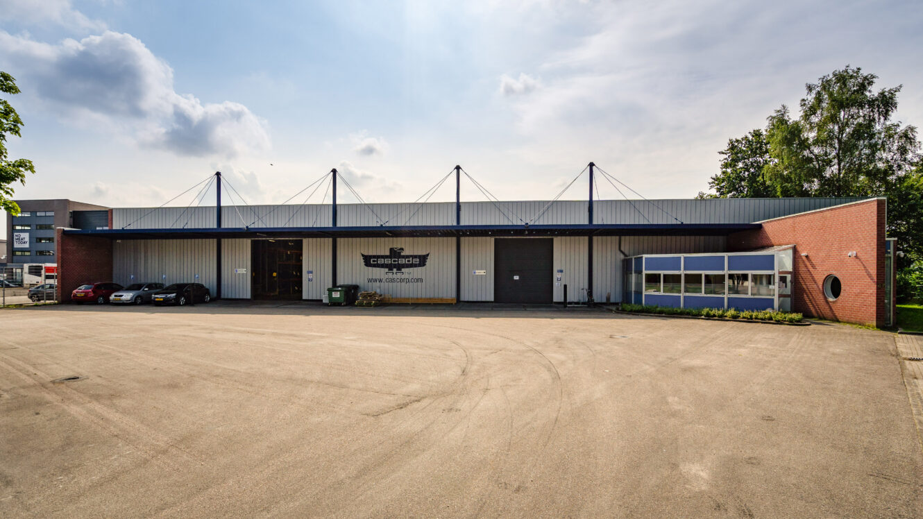 Een modern industrieel pakhuis met een metalen dak, bakstenen en metalen bekleding, een klein kantoorgedeelte, geparkeerde auto's en een grotendeels lege parkeerplaats op een bewolkte dag.