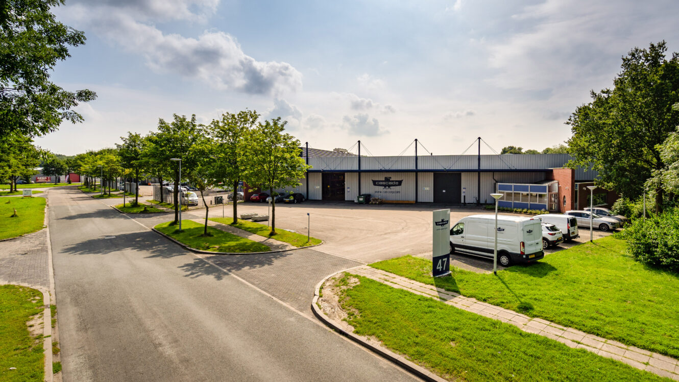 Een brede straat met geparkeerde auto's leidt naar een groot industrieel pakhuis met een bord waarop 47 staat en een aantal bomen langs het trottoir onder een gedeeltelijk bewolkte hemel.