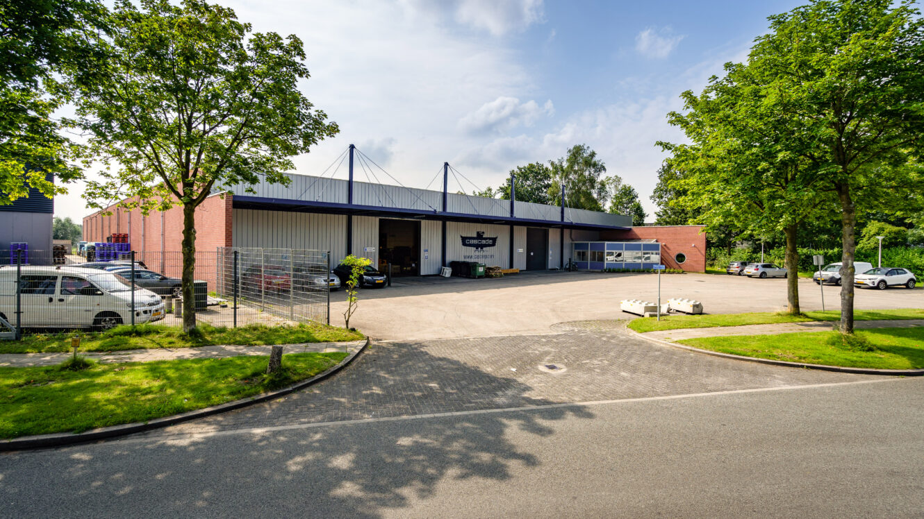 Een groot industrieel pakhuis met een omheind parkeerterrein, verschillende geparkeerde voertuigen en bomen langs het terrein onder een gedeeltelijk bewolkte hemel.