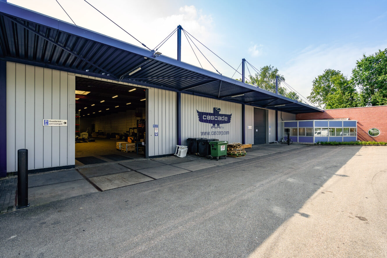 Industrieel pakhuis aan de buitenkant met grote open deuren, Cascade-bord boven de ingang, laadruimte, bakken en apparatuur zichtbaar binnen; bomen en lucht op achtergrond.