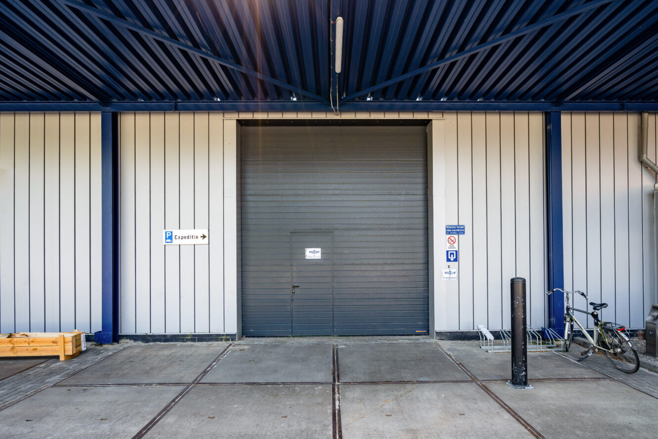 Een gesloten industriële garagedeur met borden, een fiets en een klein houten kratje vlakbij op een betonnen oppervlak.