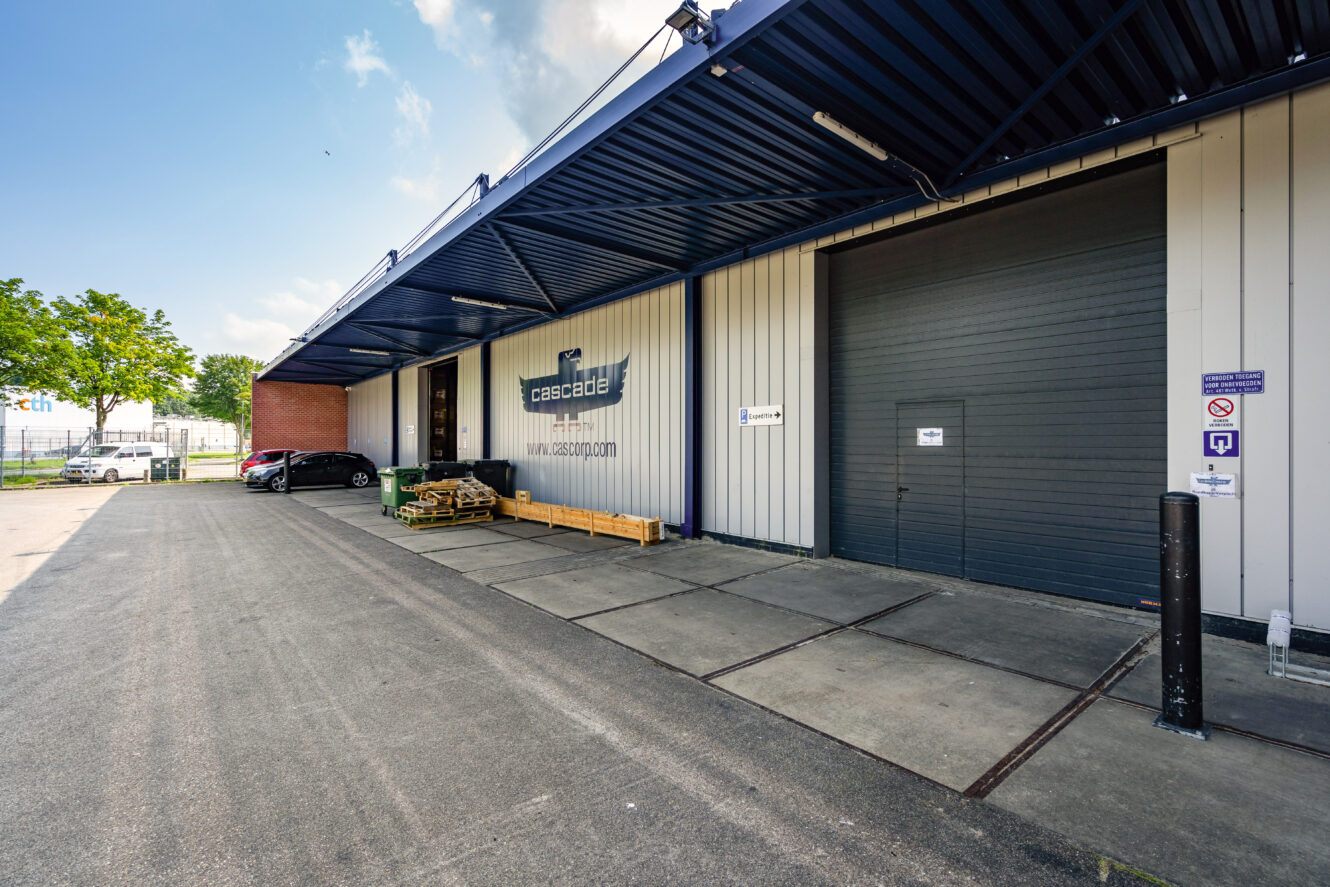 Industrieel magazijn aan de buitenkant met grote roldeur, metalen luifel en verschillende geparkeerde auto's; bedrijfslogo "Cascade" zichtbaar op de muur van het gebouw.
