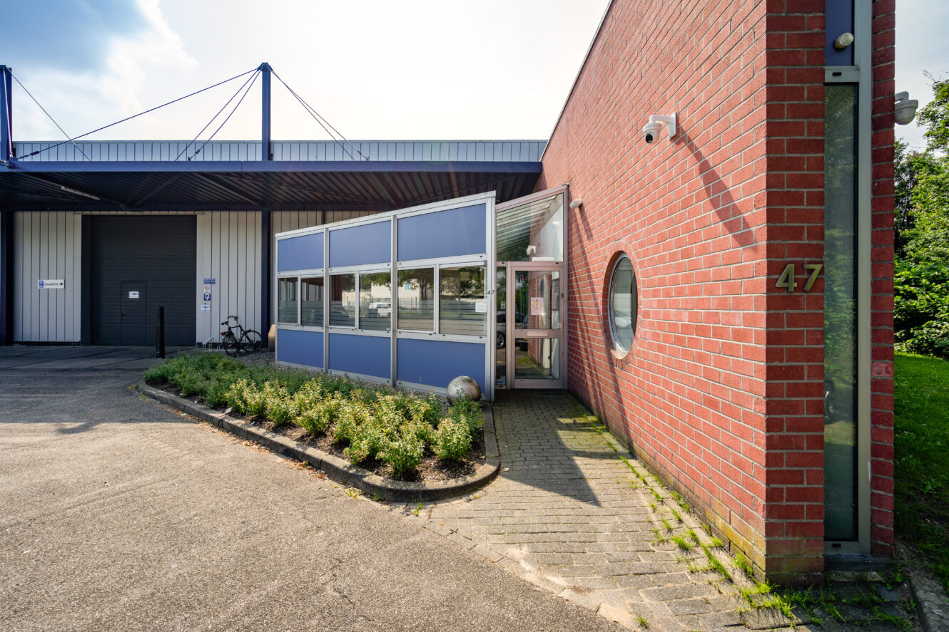 Bakstenen en metalen gebouw aan de buitenkant met glazen ingang, beveiligingscamera en groot nummer 47 op de muur. Struiken omzomen het looppad; overhang en fietsenrekken zijn zichtbaar op de achtergrond.