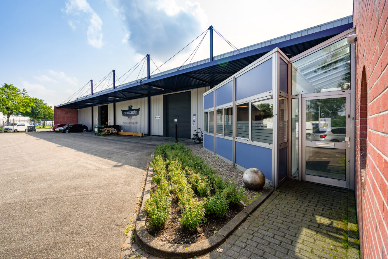 Industrieel gebouw met een blauw metalen dak, glazen ingang, geparkeerde auto's en een klein aangelegd gebied aan de voorkant op een zonnige dag.