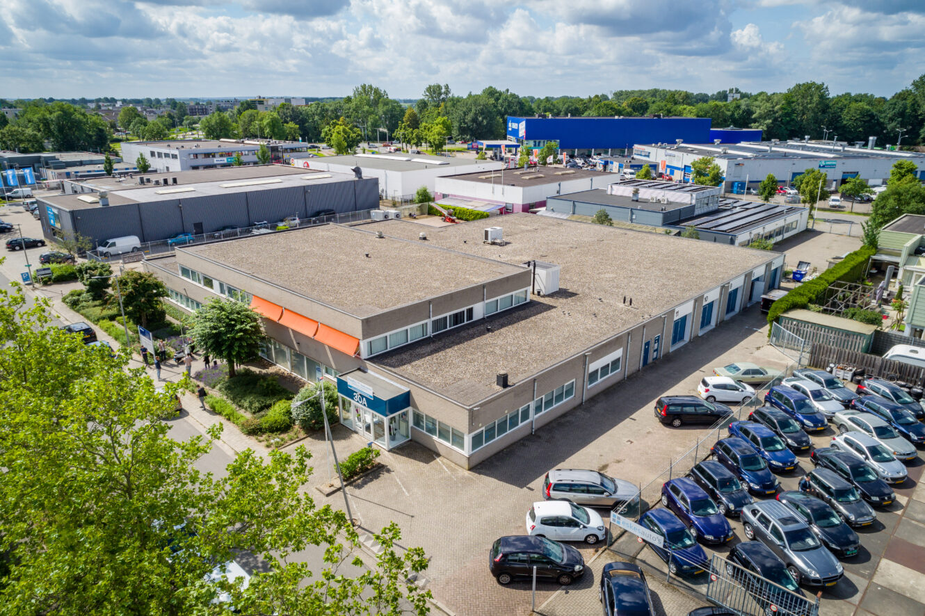 Luchtfoto van een commercieel of industrieel gebouwencomplex met een plat dak, omringd door geparkeerde auto's en nabijgelegen magazijnen, onder een gedeeltelijk bewolkte hemel.