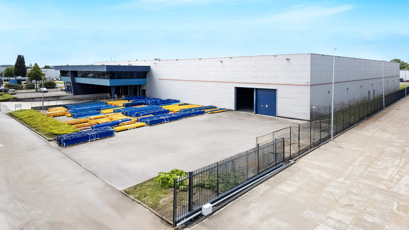 Groot industrieel pakhuis met een blauwe garagedeur, omheinde binnenplaats en stapels blauwe en gele metalen buizen die buiten op het betonnen terrein staan.