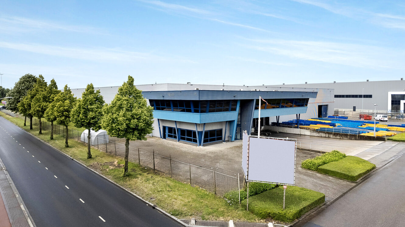 Een modern industrieel gebouw met blauwe accenten is omringd door bomen en hekwerk, gelegen naast een weg met een leeg bord ervoor.