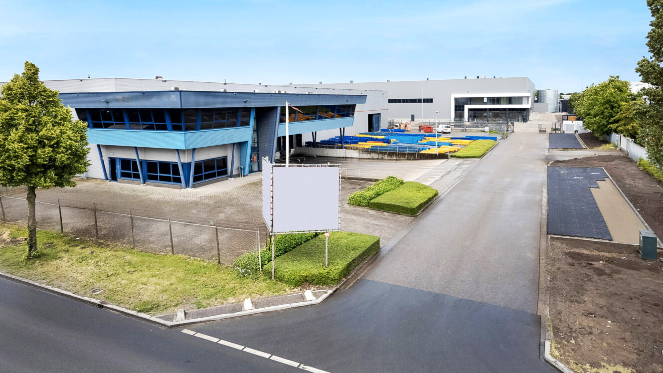 Industriële faciliteit met een groot grijs magazijn, een gebouw met blauwe randen, een parkeerplaats, aangelegde struiken en een leeg wit bord bij de ingang onder een heldere hemel.