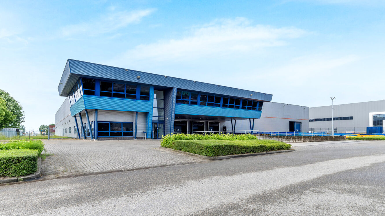 Modern industrieel pakhuis met blauwe accenten, grote ramen en een verharde oprit omringd door gesnoeide heggen onder een heldere hemel.