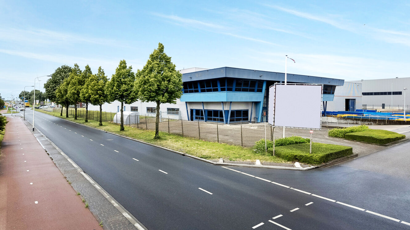 Een modern industrieel gebouw met blauwe accenten staat naast een lege straat met bomen; een groot leeg reclamebord staat voor het pand.