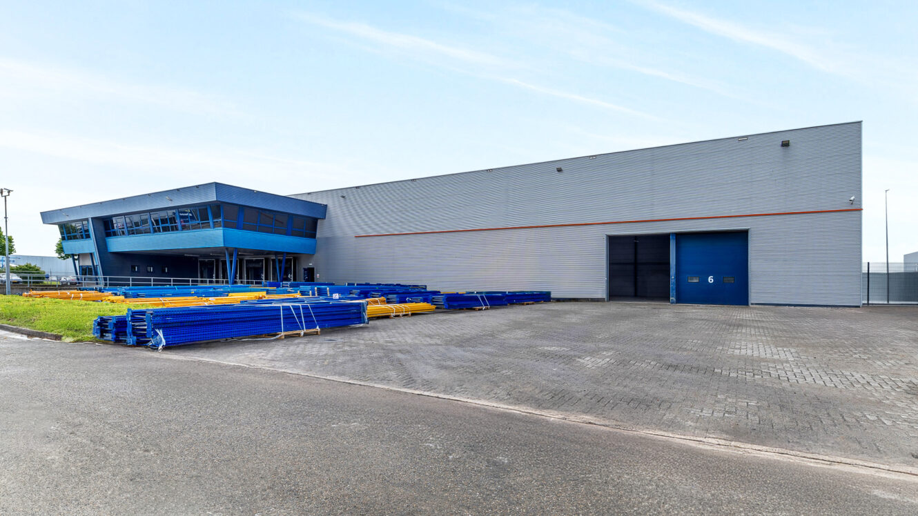 Een groot industrieel magazijn met een blauwe en grijze buitenkant, aangrenzend kantoorgedeelte en stapels blauwe en gele materialen buiten op een verhard terrein.