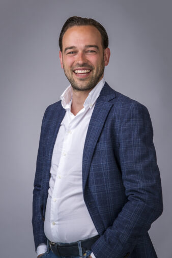 Een man in een blauw geruit colbert en wit overhemd staat te glimlachen tegen een effen grijze achtergrond.