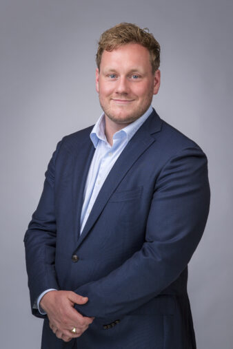 Man in marineblauw pak en lichtblauw overhemd poseert voor een professioneel portret tegen een effen grijze achtergrond.