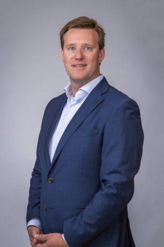 Een man in een blauw pak en wit overhemd staat tegen een effen, lichtgrijze achtergrond en kijkt met een neutrale uitdrukking naar de camera.