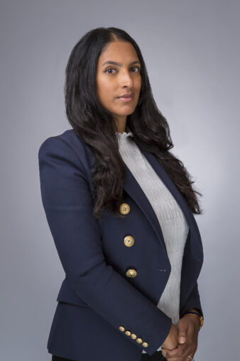 Een vrouw met lang donker haar, draagt een marineblazer met gouden knopen en een lichte blouse, staat met haar gezicht naar de camera tegen een effen grijze achtergrond.