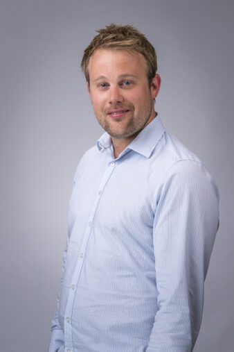 Een man met kort lichtbruin haar en een getrimde baard draagt een lichtblauw overhemd met knoopsluiting, staande voor een effen grijze achtergrond.