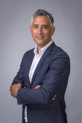 Man in een marineblauw pak en wit overhemd staat met gekruiste armen te glimlachen tegen een effen lichtgrijze achtergrond.