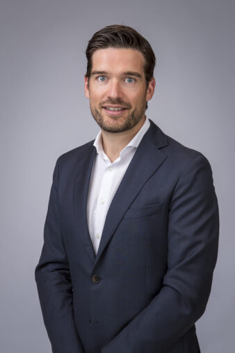 Een man met kort bruin haar en een baard in een donker pak en wit overhemd staat tegen een effen grijze achtergrond, met zijn gezicht naar de camera.