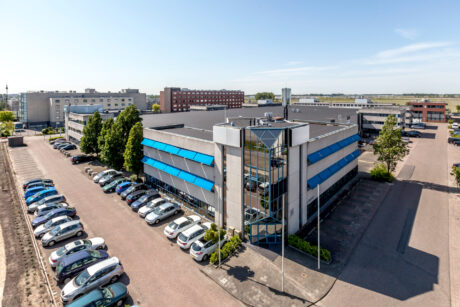 Een modern kantoorgebouw met grote ramen en blauwe luifels, omringd door geparkeerde auto's op een parkeerplaats, met andere kantoorgebouwen op de achtergrond op een heldere dag.