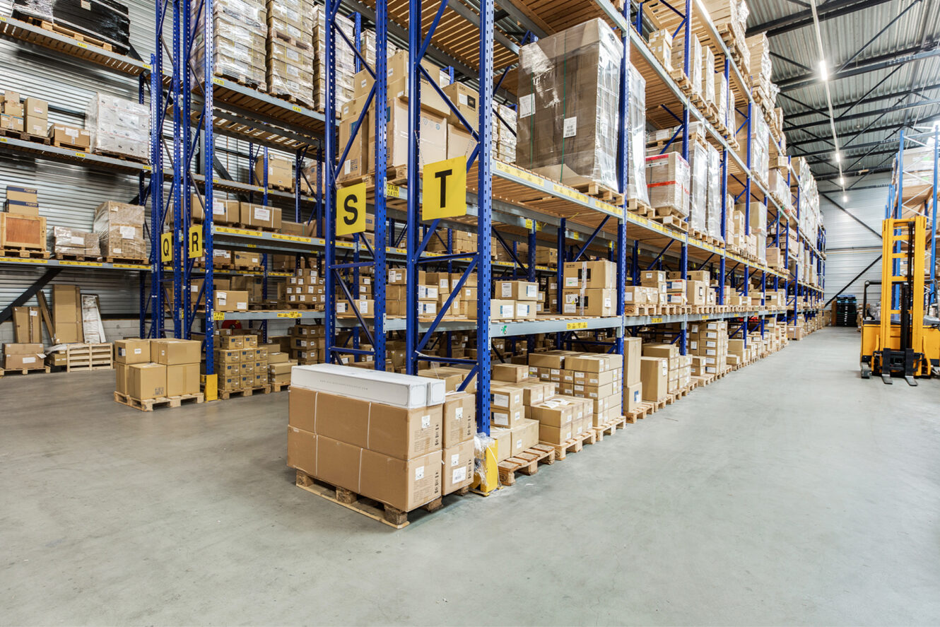 Rijen rekken in een groot magazijn, gevuld met gestapelde kartonnen dozen en pallets, met industriële rekken met letters.