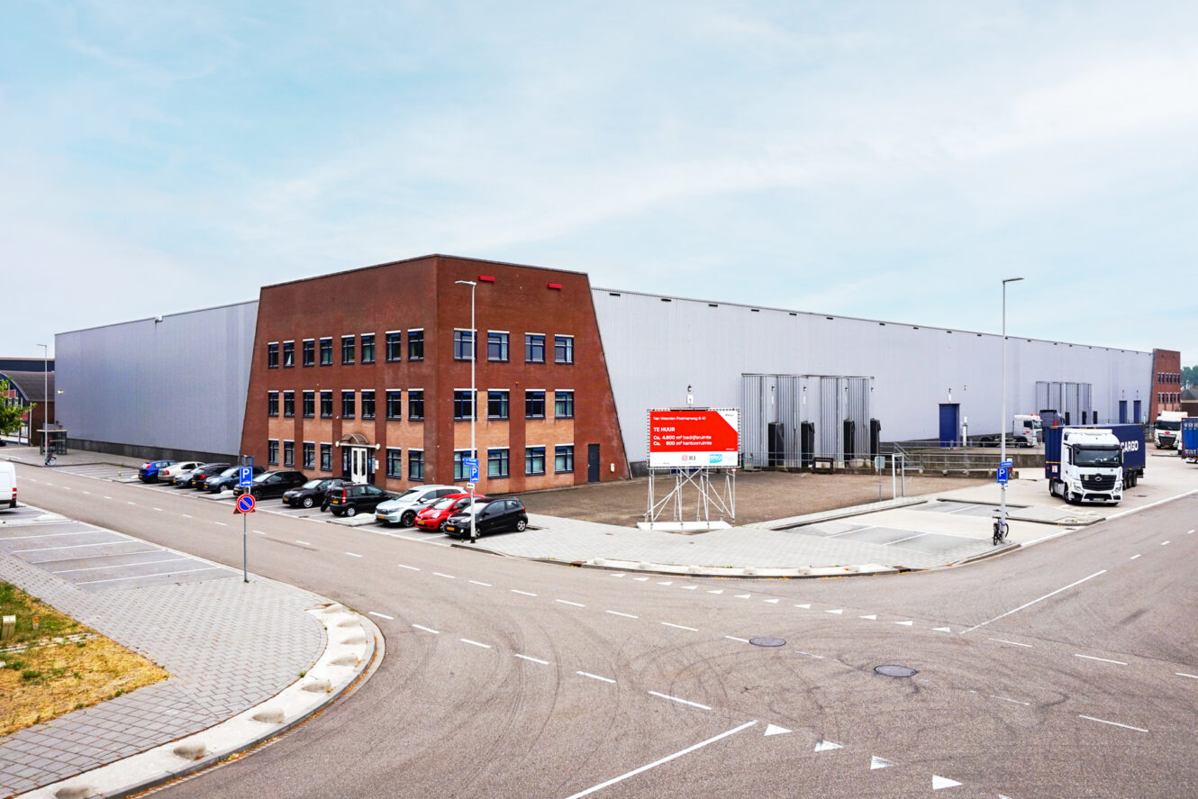 Groot industrieel pakhuis met een bakstenen kantoorgedeelte, omringd door geparkeerde auto's en vrachtwagens, onder een heldere hemel.