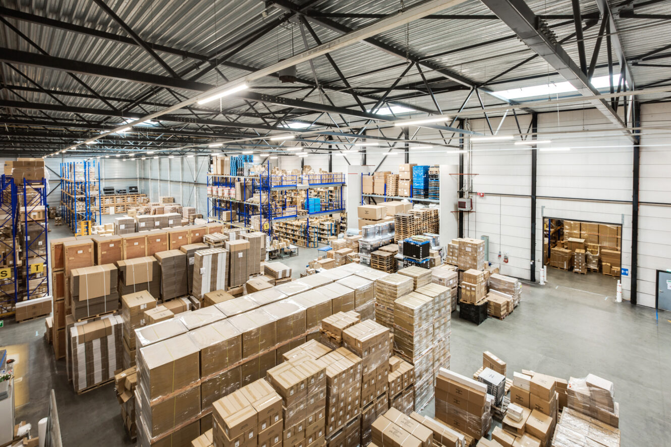 Groot magazijninterieur gevuld met gestapelde kartonnen dozen en planken, met hoge plafonds en industriële verlichting.