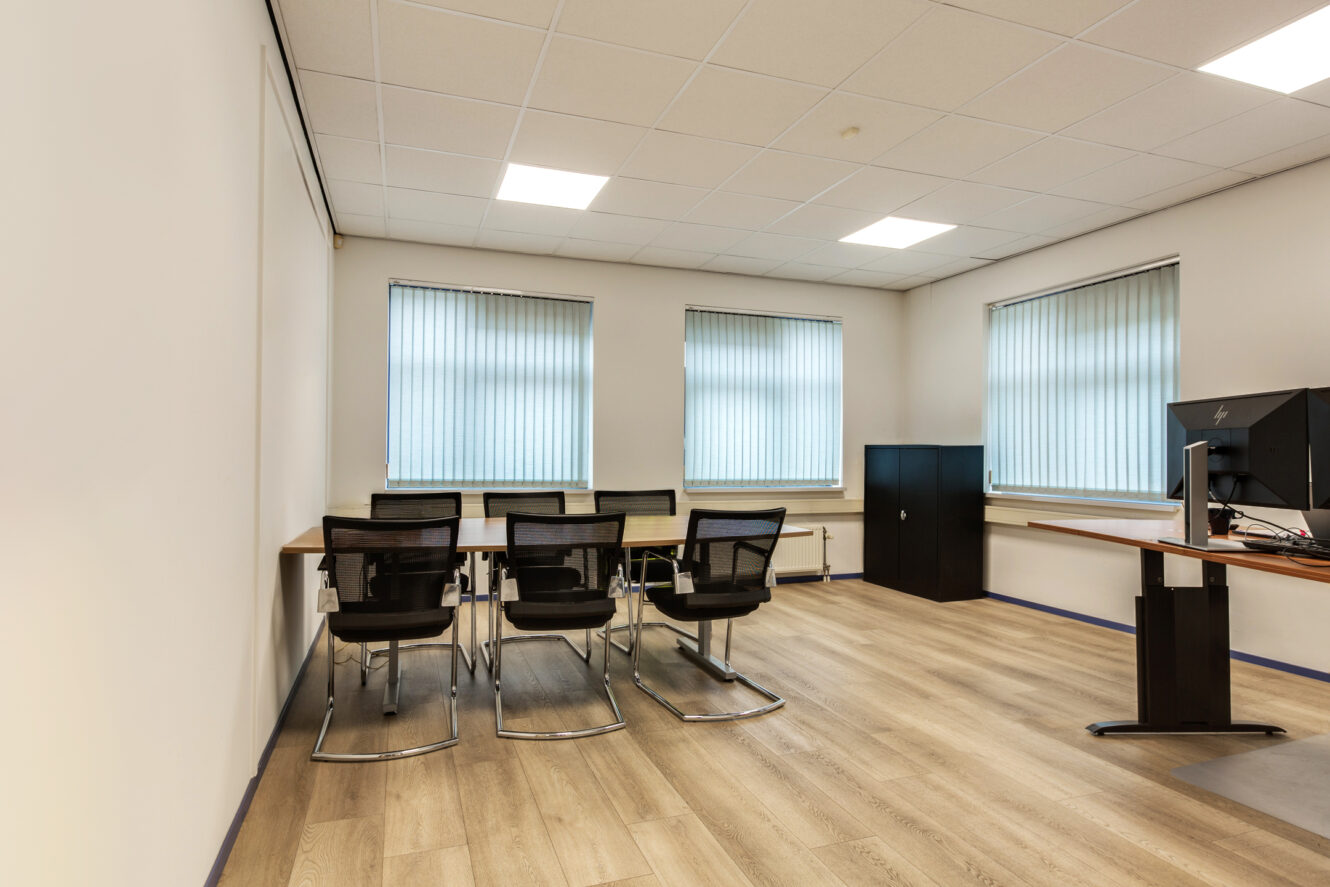 Modern kantoor met een houten vloer, drie ramen met luxaflex, een rij van vier zwarte stoelen aan een vergadertafel en een bureau met een computermonitor aan de rechterkant.
