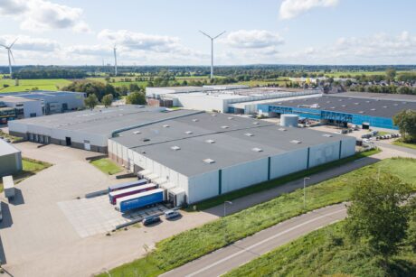 Luchtfoto van een groot industrieel magazijn met laadkades, buiten geparkeerde vrachtwagens en windturbines op de achtergrond, omringd door groen en andere commerciële gebouwen.