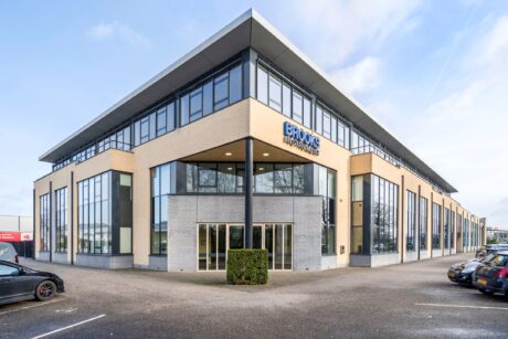 Modern kantoorgebouw van twee verdiepingen met grote ramen en een bord BROOKS op de gevel, omringd door geparkeerde auto's en een kleine haag bij de ingang.