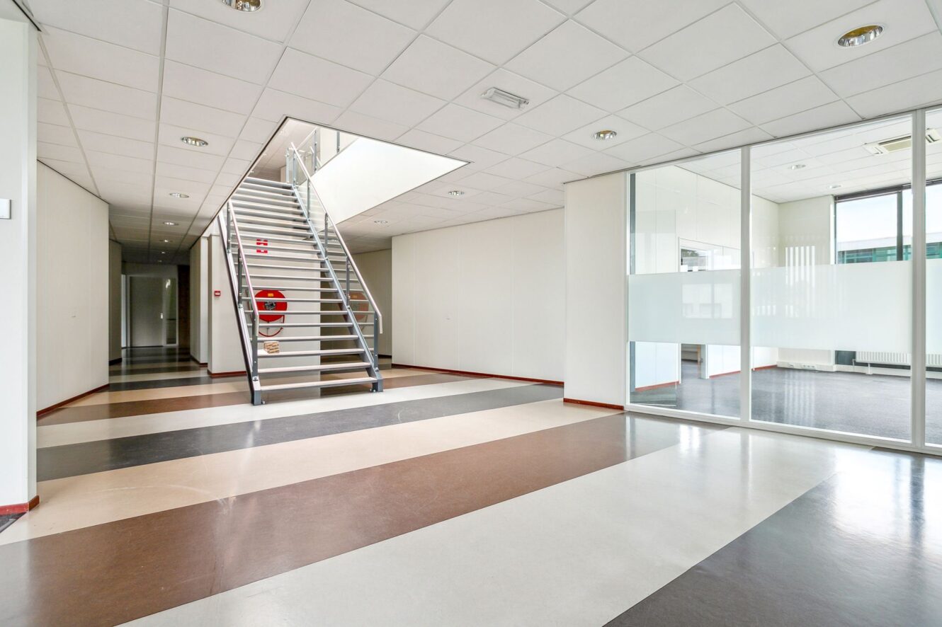 Modern kantoorinterieur met gestreepte vloeren, witte muren, een glazen scheidingswand en een metalen trap naar een bovenverdieping. Aan de muur bij de trap is een rode brandslanghaspel bevestigd.