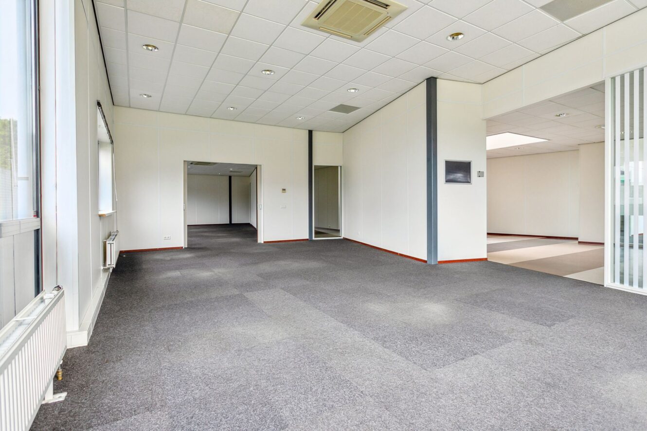Lege kantoorruimte met grijs tapijt, witte muren, grote ramen aan de linkerkant en plafondverlichting. Gedeelde ruimtes en open deuropeningen leiden naar aangrenzende kamers.