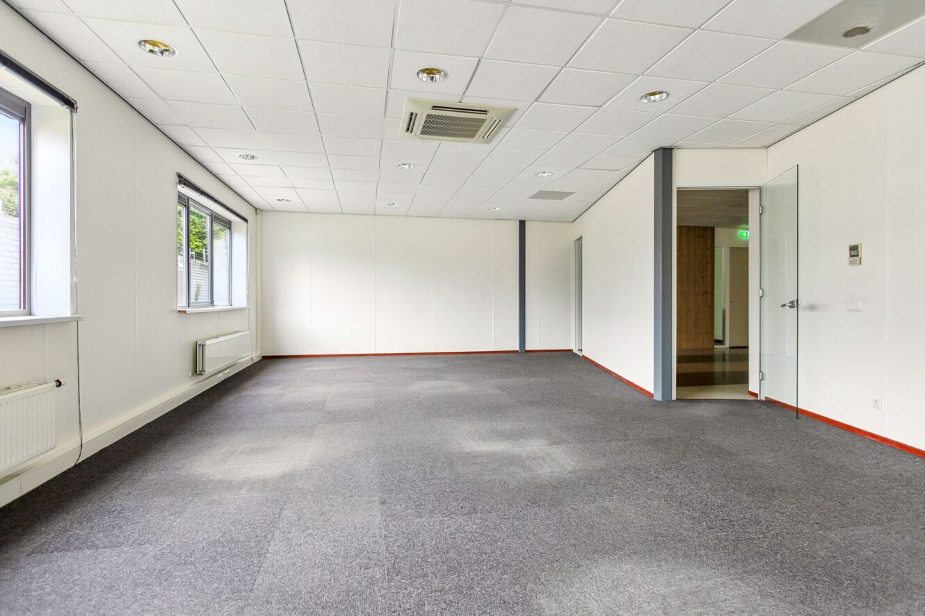 Lege kantoorkamer met grijs tapijt, witte muren, plafondverlichting, grote ramen aan de linkerkant en een open glazen deur die naar een gang leidt.