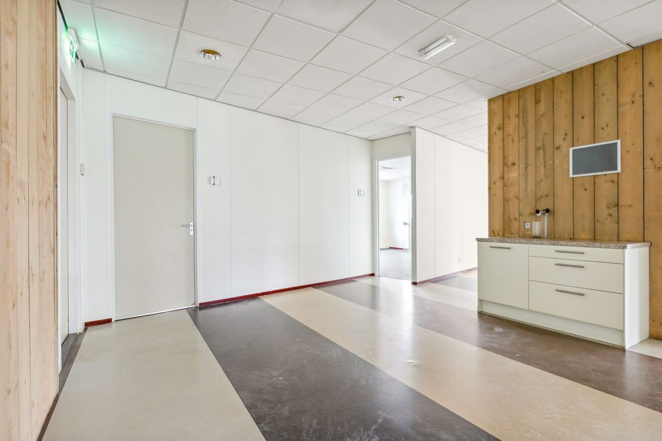 Een moderne, lege kamer met een vloer met patroon, witte muren, een accentmuur met houten panelen, een klein keukentje en verschillende deuropeningen.