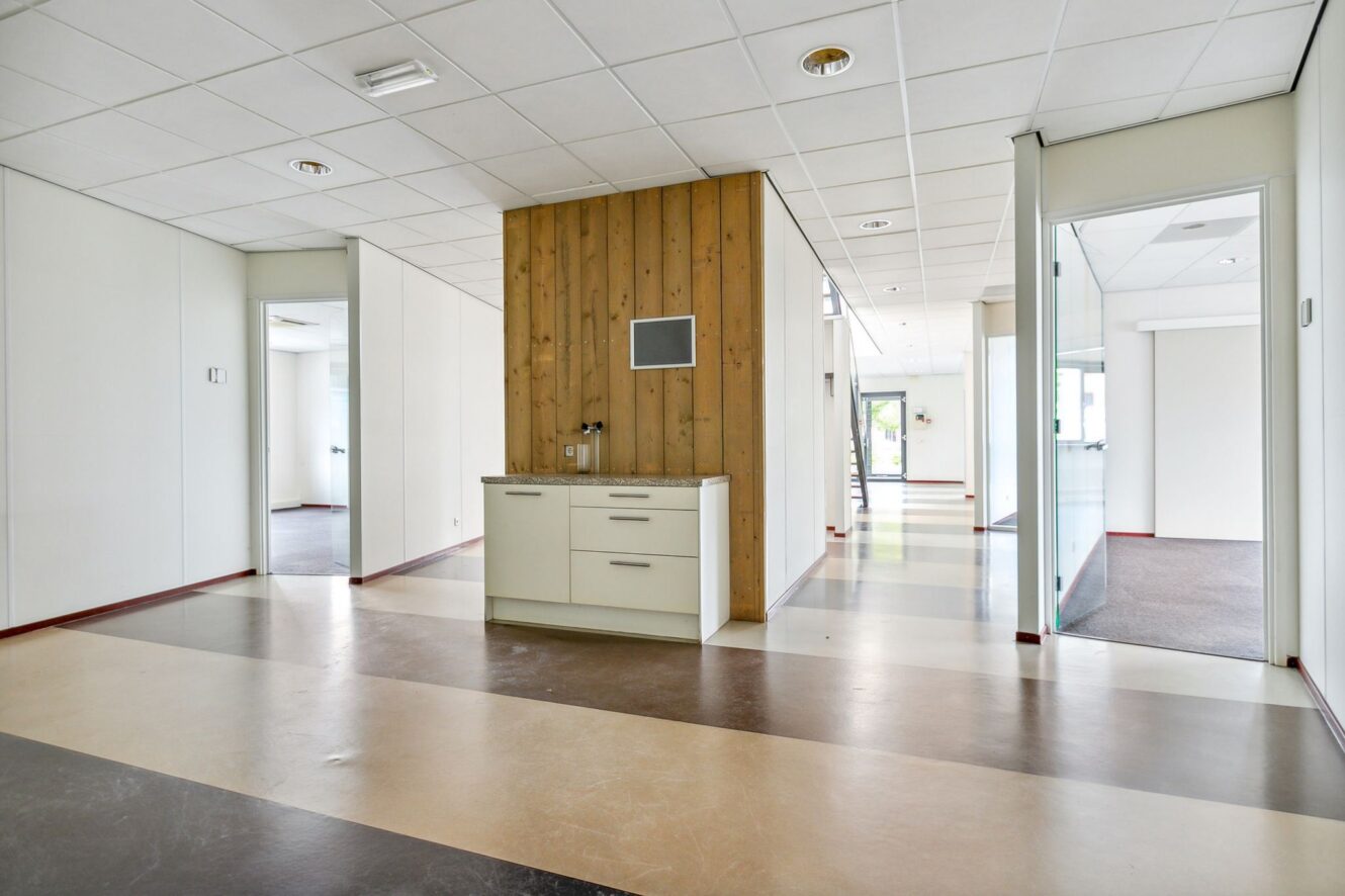 Een moderne, lege kantoorruimte met witte muren, grote ramen, glazen deuren en een klein keukentje tegen een schot met houten panelen.