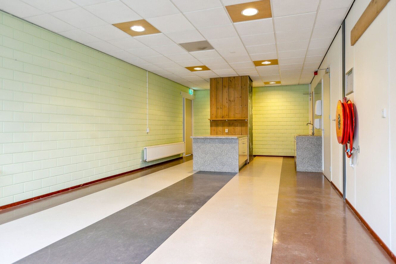 Een lange, lege kamer met betegelde vloeren, lichtgroene bakstenen muren, een kleine keuken aan het eind en een brandslanghaspel aan de muur.