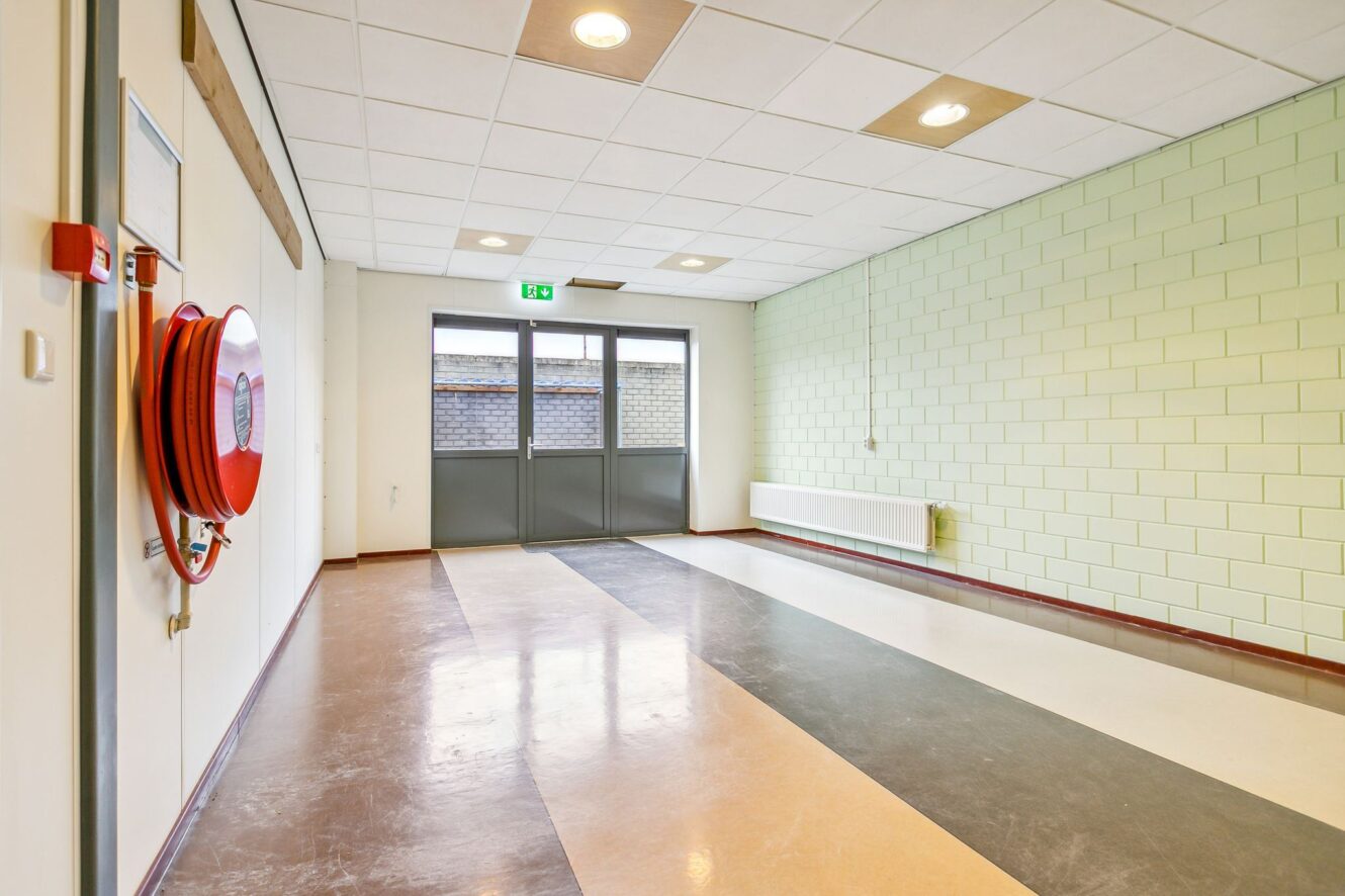Lege hal met beige, bruine en grijze vloerstrepen, een lichtgroene betegelde muur, plafondverlichting, dubbele uitgangsdeuren, een radiator en een rode brandslanghaspel aan de muur.