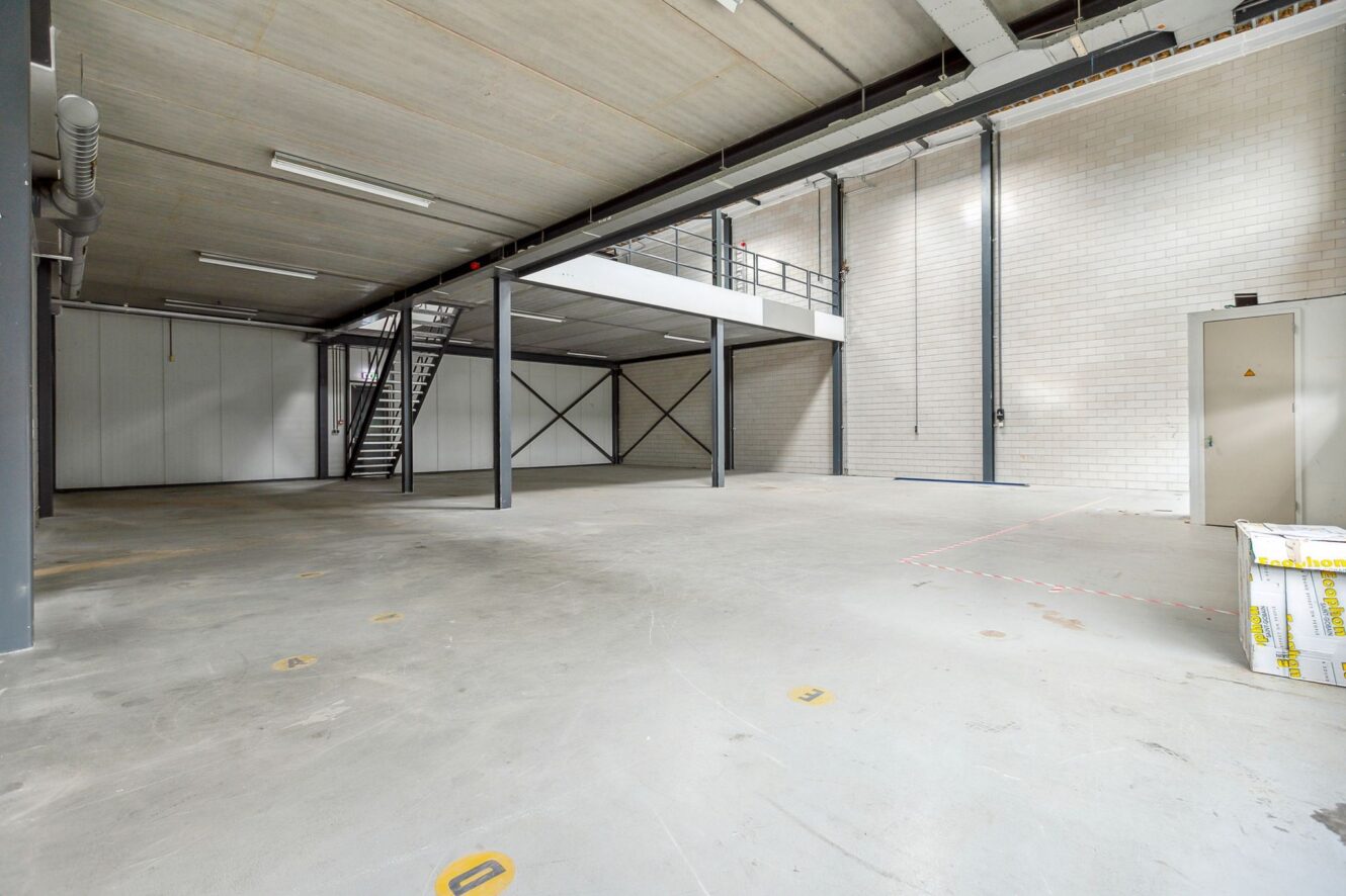 Ruim, leeg industrieel pakhuis met betonnen vloeren, hoog plafond, tussenverdieping, metalen trap en zichtbare leidingen.