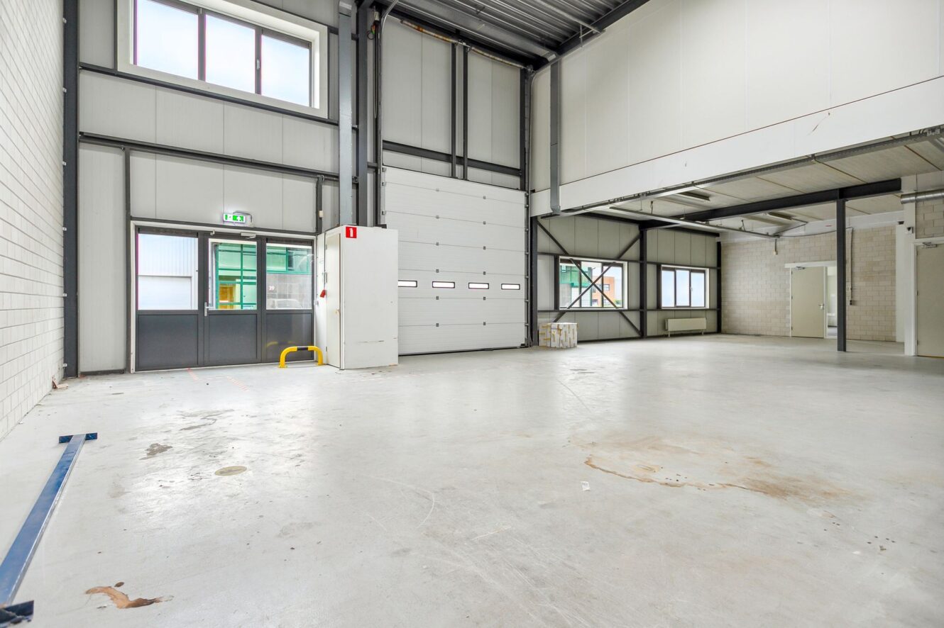 Grote, lege industriële opslagruimte met hoge plafonds, witte muren, zichtbare stalen balken en meerdere ramen en deuren.