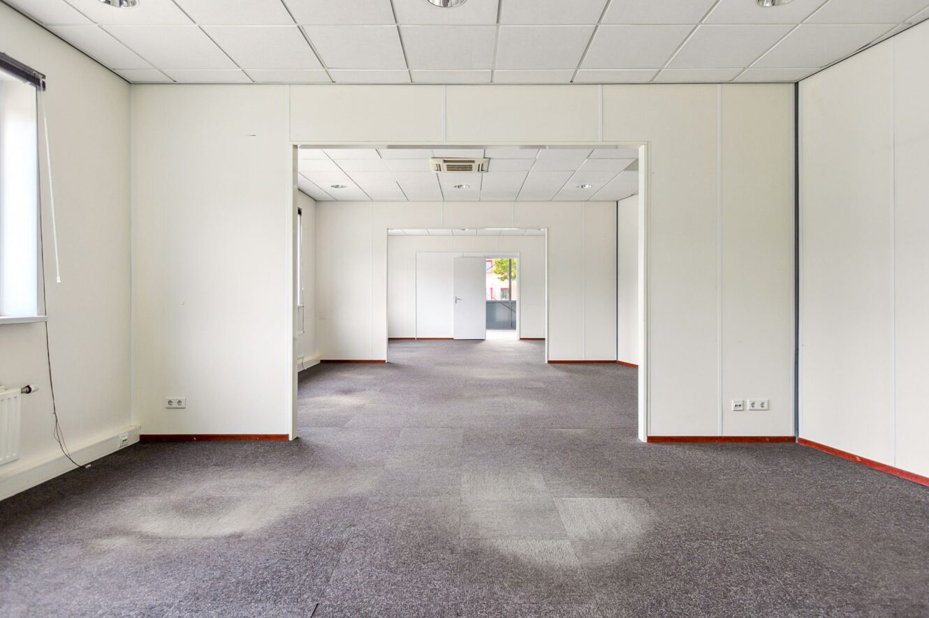 Lege kantoorruimte met witte muren, grijs tapijt, plafondverlichting en meerdere deuropeningen naar aangrenzende kamers. Natuurlijk licht valt binnen door een raam aan de linkerkant.