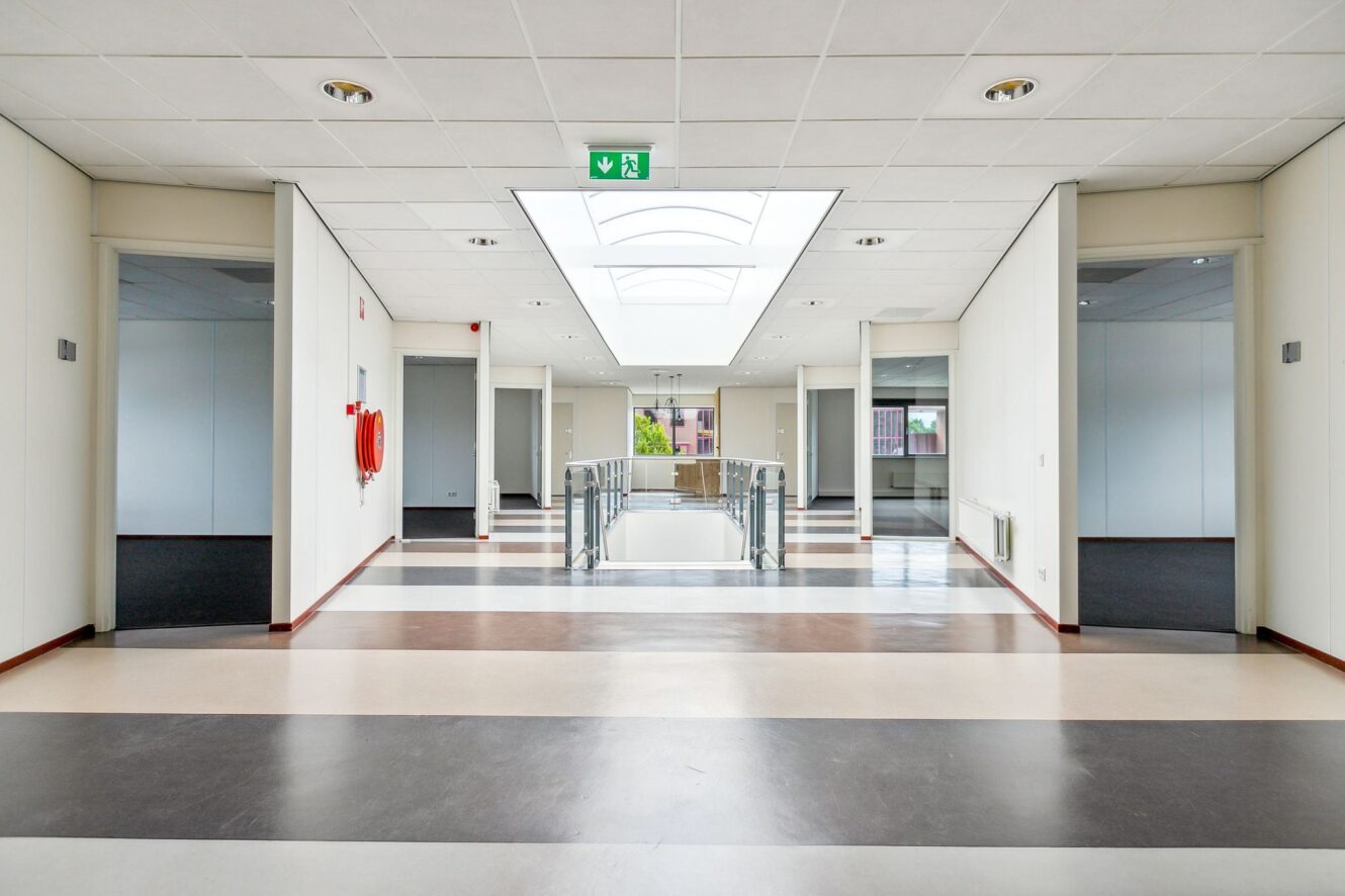 Een moderne, lege gang met gestreepte vloeren, witte muren, brandblussers en een bord met de nooduitgang aan het plafond. Meerdere deuropeningen omzomen de gang.