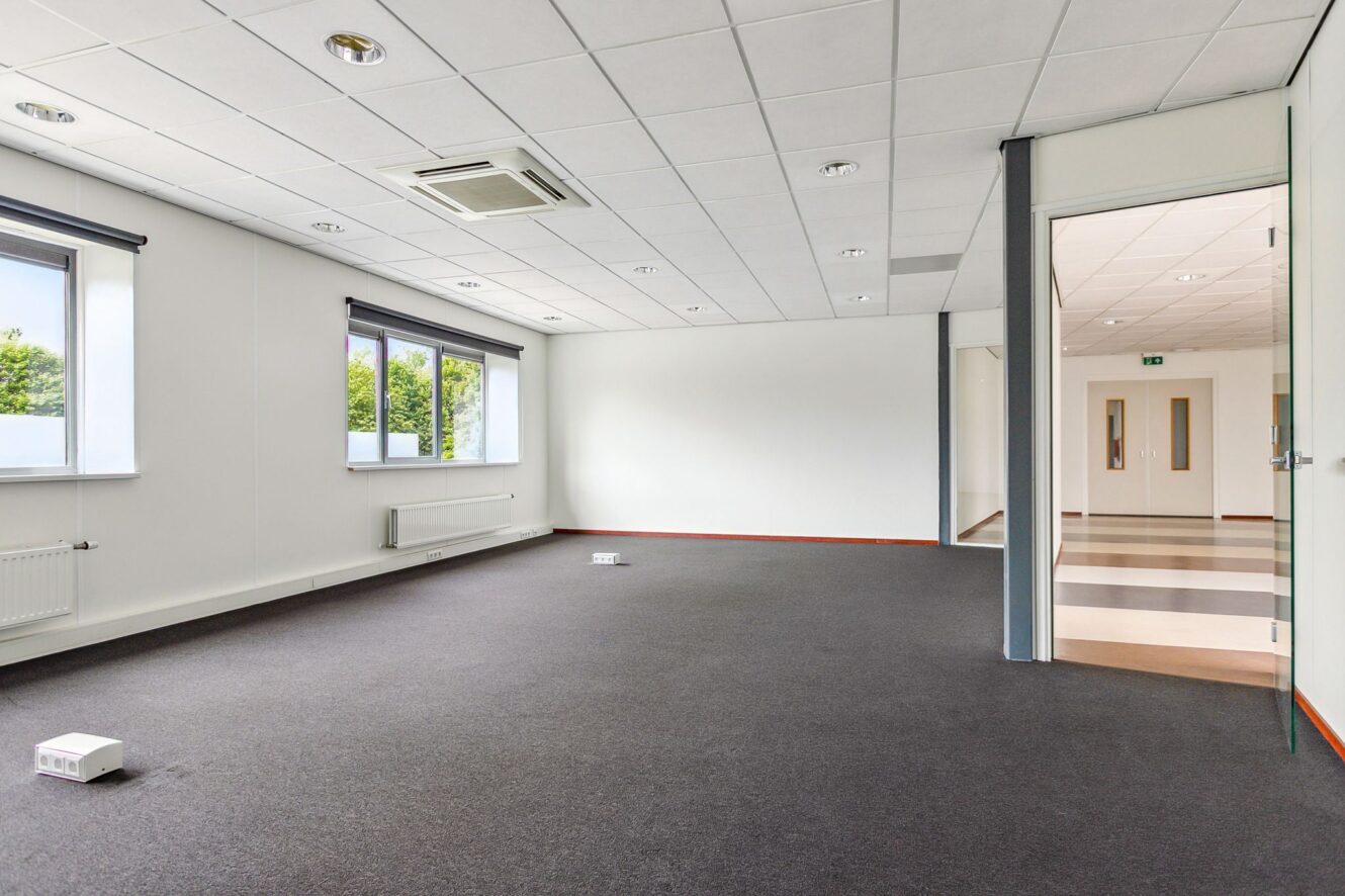 Lege kantoorkamer met witte muren, grijs tapijt, grote ramen, plafondverlichting en een open deur die naar een hal leidt.