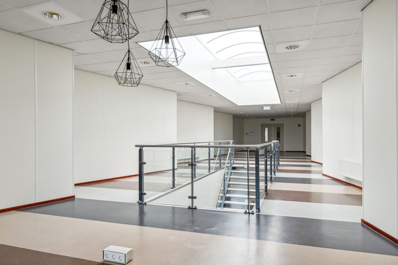 Moderne hal binnenshuis met een dakraam, geometrische hanglampen, glazen balustrade en een trap die naar een lager niveau leidt. De vloer heeft afwisselend bruine, beige en zwarte strepen.