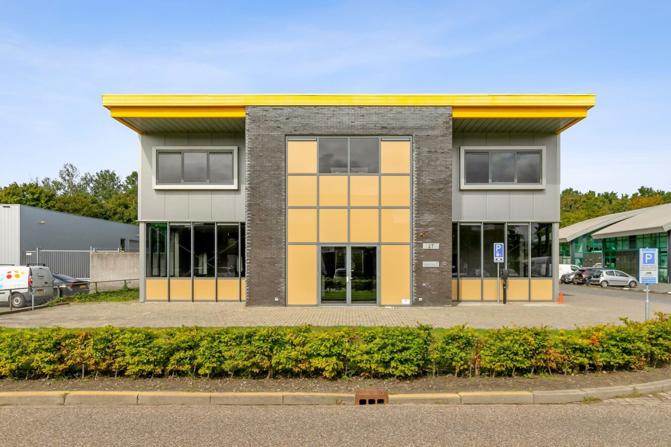 Modern commercieel gebouw van twee verdiepingen met gele accenten, grote ramen en een geplaveide parkeerplaats aan de voorkant, omzoomd door lage struiken.