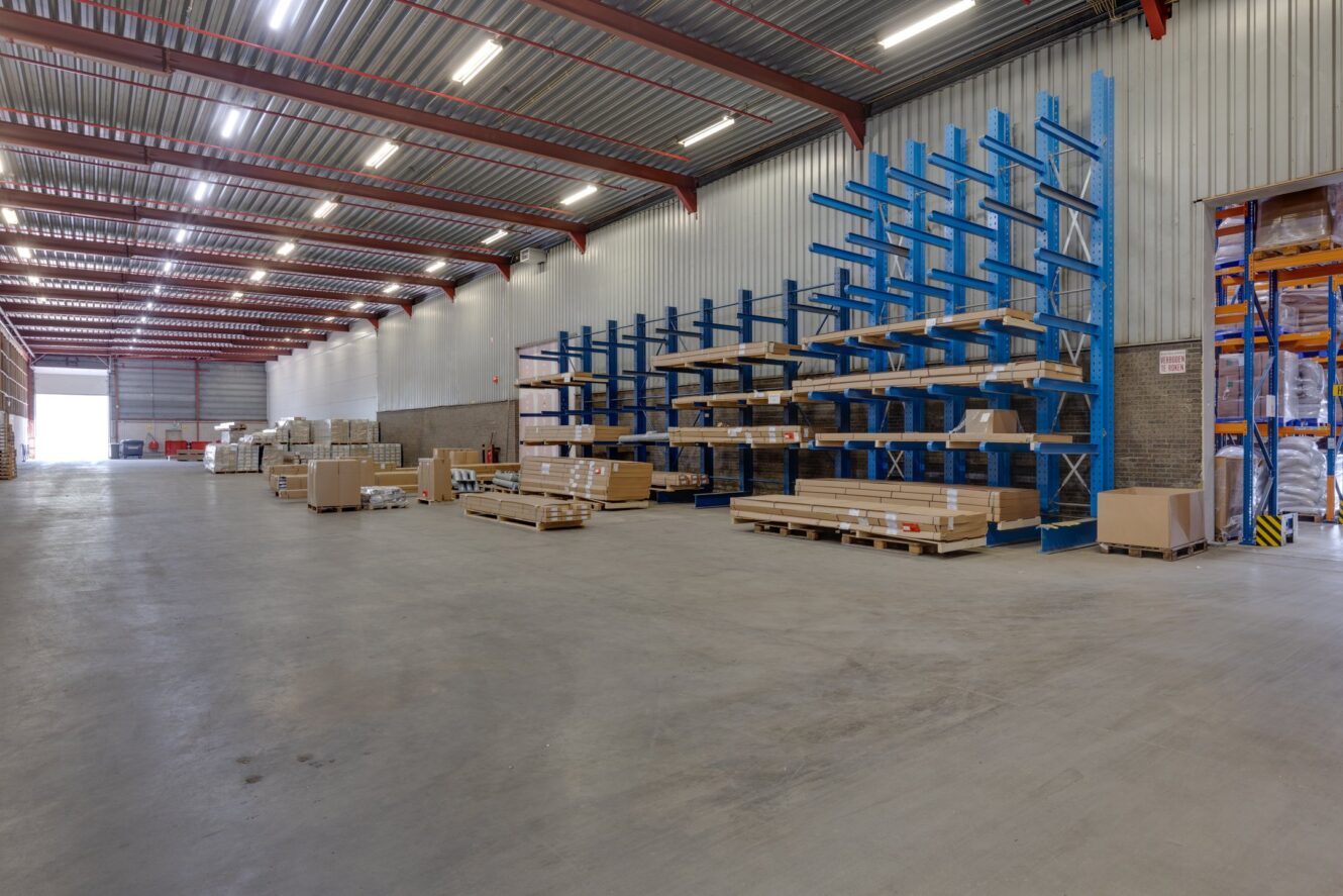 Ruim magazijninterieur met hoge plafonds, metalen planken met pallets en dozen en gestapelde goederen die langs de muren zijn georganiseerd.