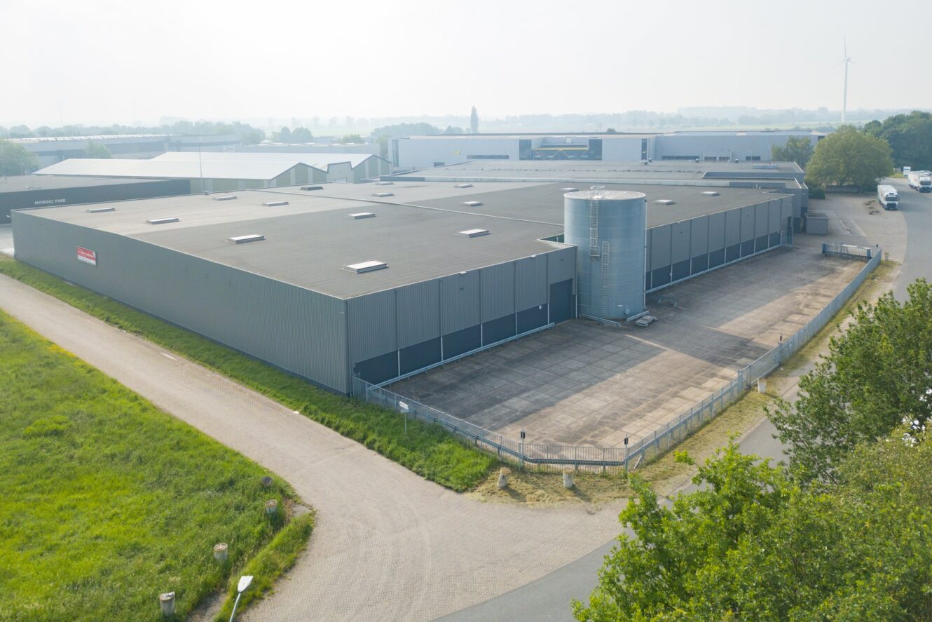 Een groot, grijs industrieel pakhuis met een cilindrische metalen silo, omgeven door een verhard terrein en omheind terrein, gelegen in de buurt van een weg en grasvelden.